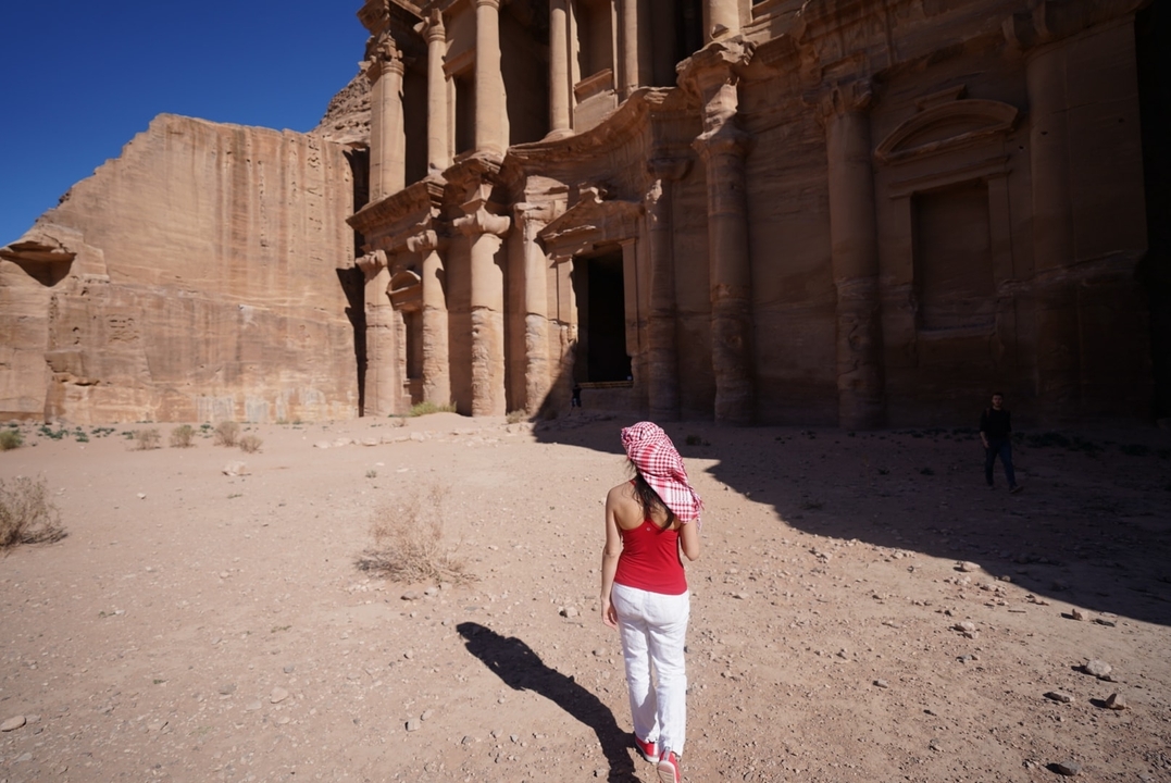 Woman in red top walks toward the imposing rock-carved Monastery at Petra under bright desert sun.