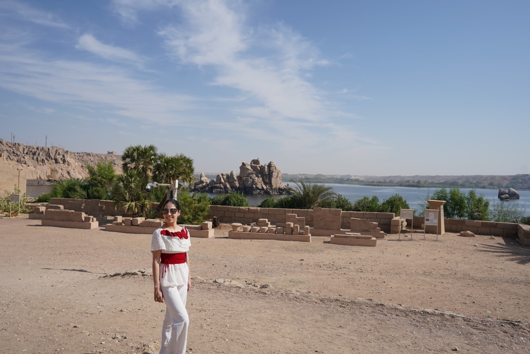 Traveler in white and red outfit stands by ancient riverside ruins with palm trees and Nile waters beyond.