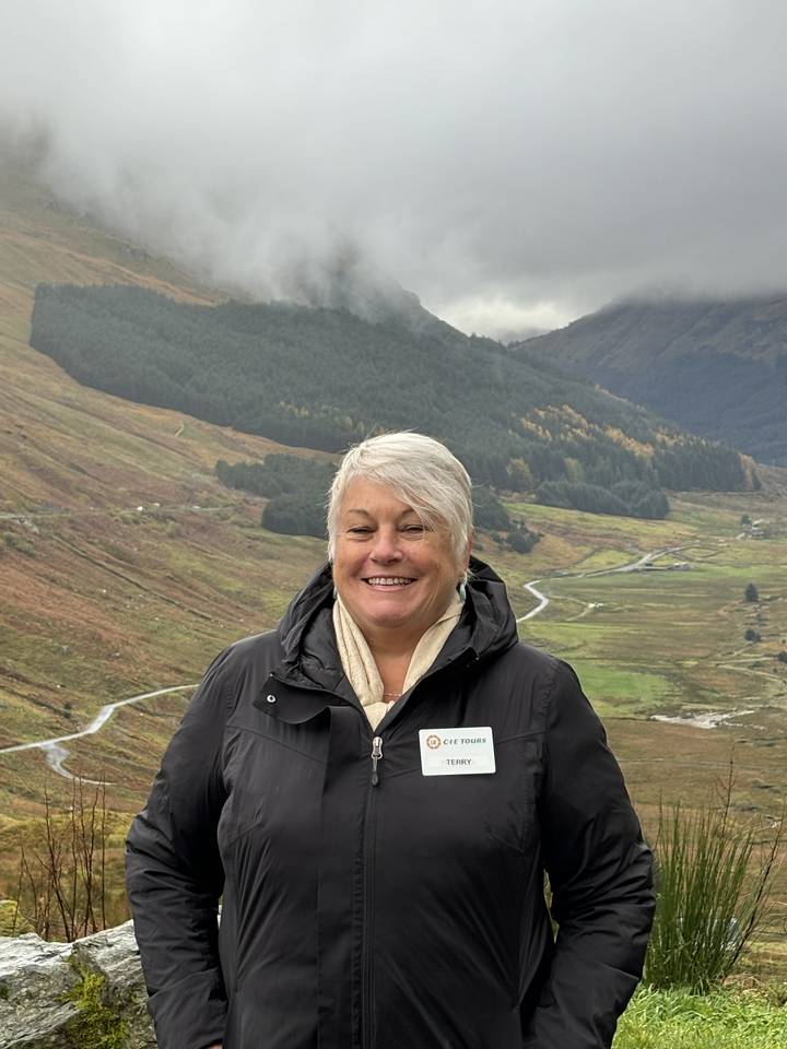 Une personne qui pose dans une campagne montagneuse avec un ciel nuageux.