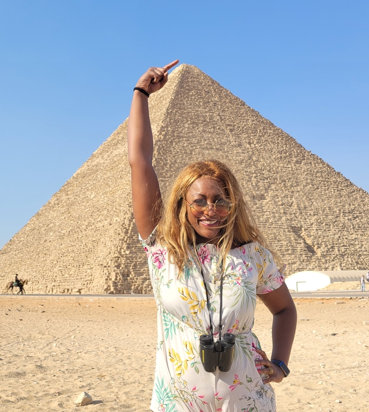 Personne posant avec une pyramide en arrière-plan sous un ciel bleu dégagé.