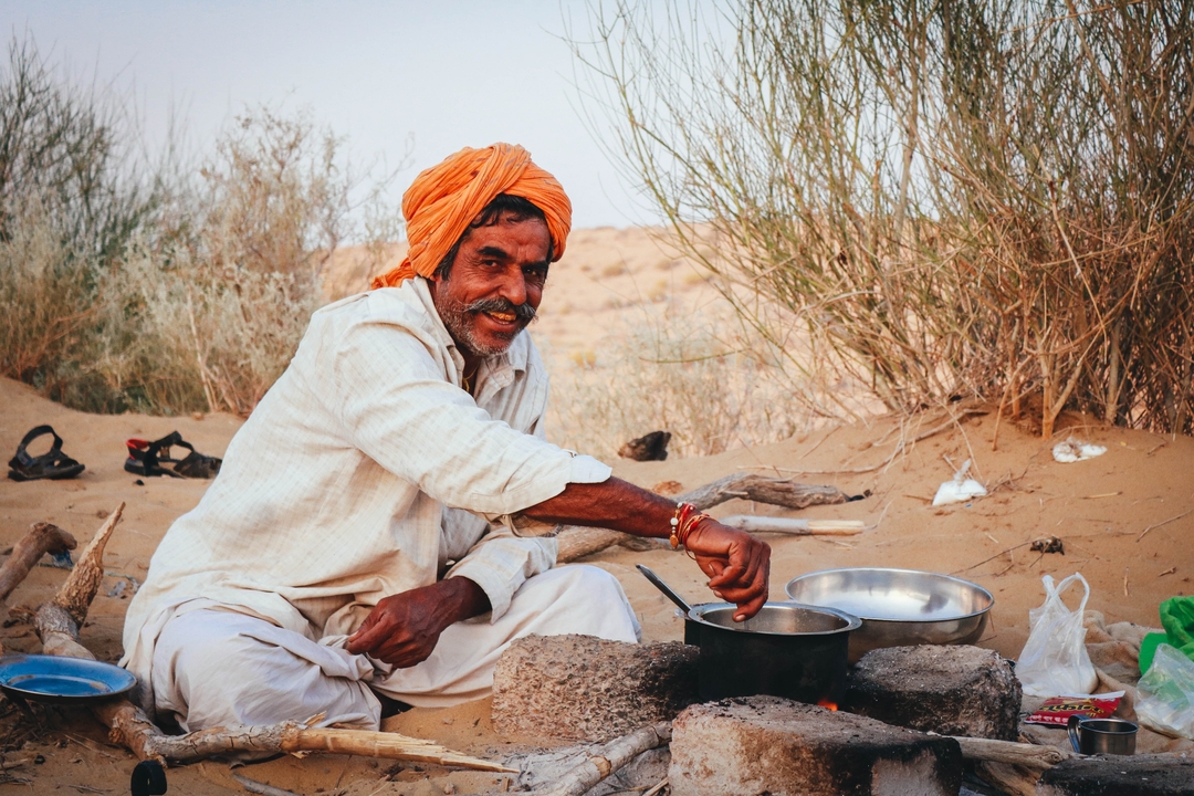 Rajasthani man in turban smiles while cooking over a small campfire amid golden desert sand and scrub.