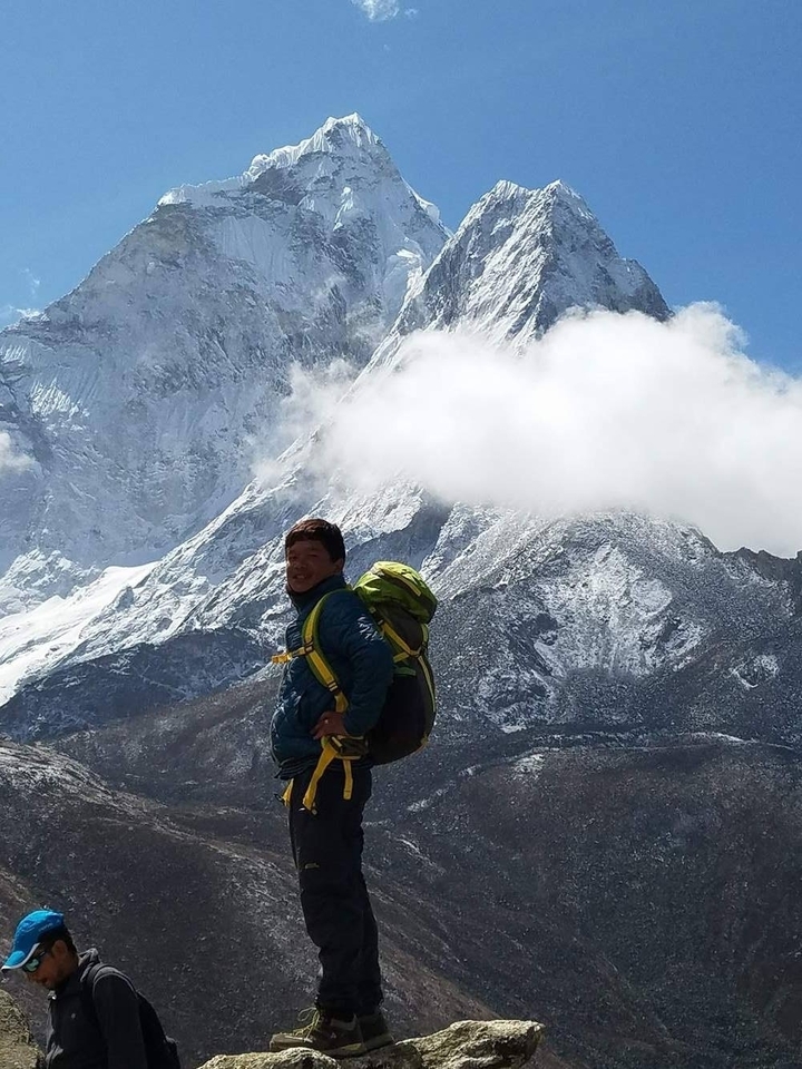 Smiling trekker with green backpack poses before a towering snow-covered Himalayan peak and drifting cloud.