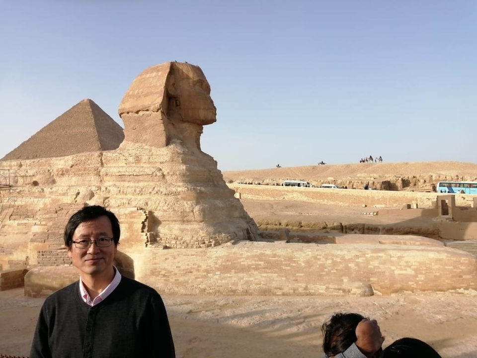 Tourist standing before the Great Sphinx with the Pyramid of Khafre rising behind under a hazy desert sky.