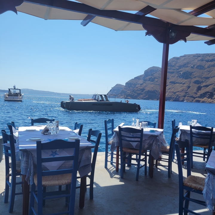 Tables de salle à manger dressées au bord de la mer avec des bateaux et des montagnes en arrière-plan.