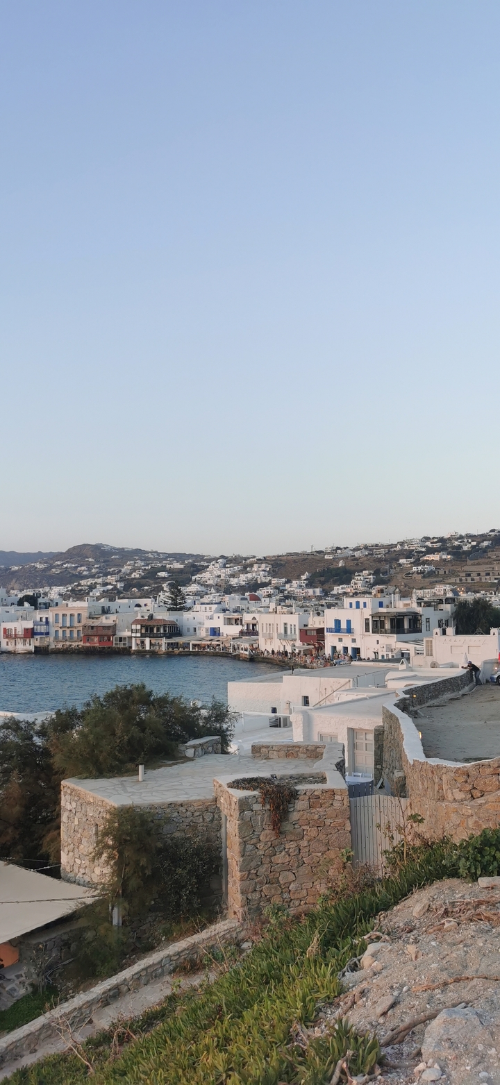 Daytime panorama of whitewashed buildings spilling down the hillsides of Mykonos beside blue water.