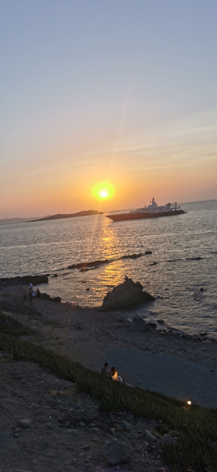 Golden sunset over the Aegean Sea with a luxury yacht silhouetted against shimmering water near rocky shores.