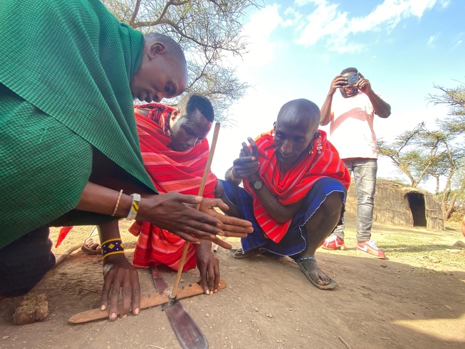 Maasai men demonstrate traditional fire-making by rubbing sticks together while a visitor photographs.
