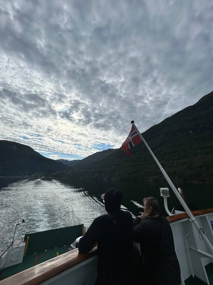 Persons on a boat deck with a flag and fjord view.