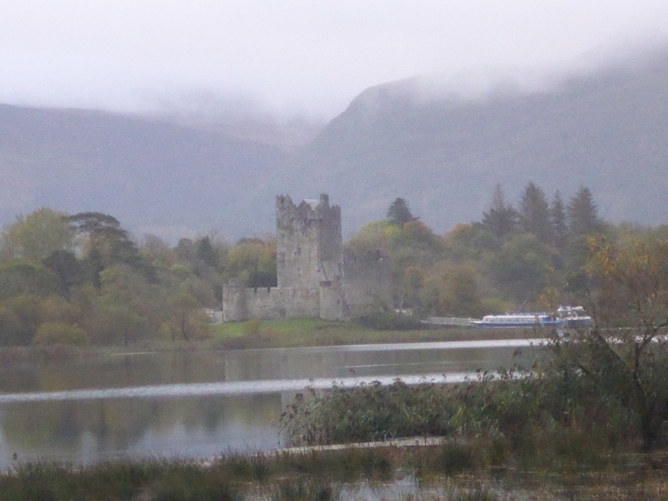 Schloss mit einem Boot auf einem See, umgeben von Bäumen und Hügeln.