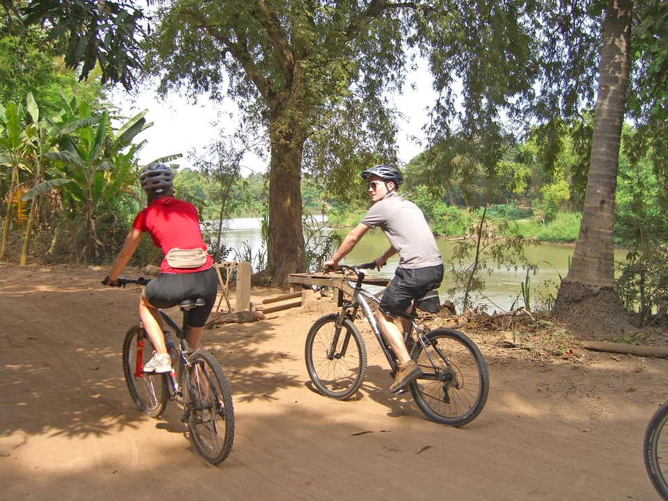 Deux personnes faisant du vélo le long d'un sentier de terre à côté d'une rivière avec des arbres.