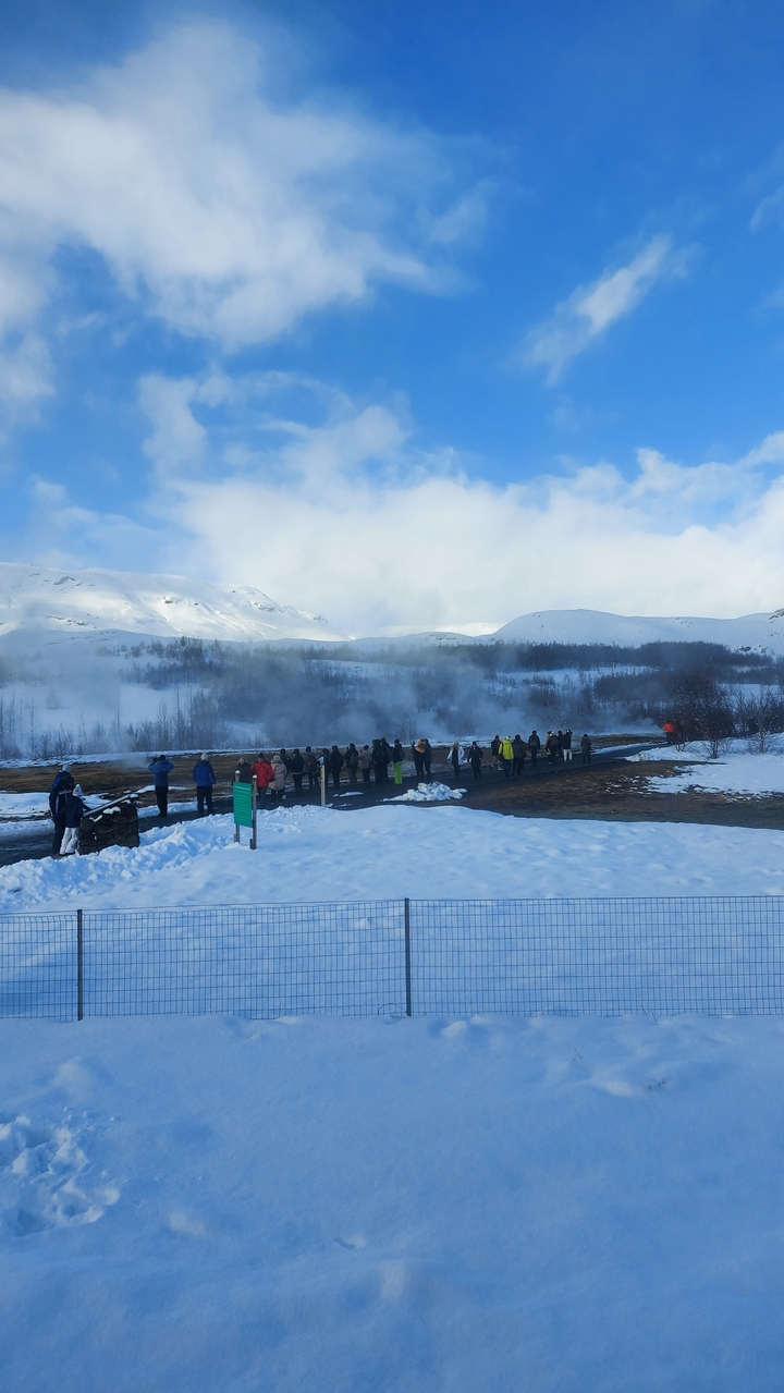 Des touristes visitant une zone de geysers avec des montagnes enneigées en arrière-plan.