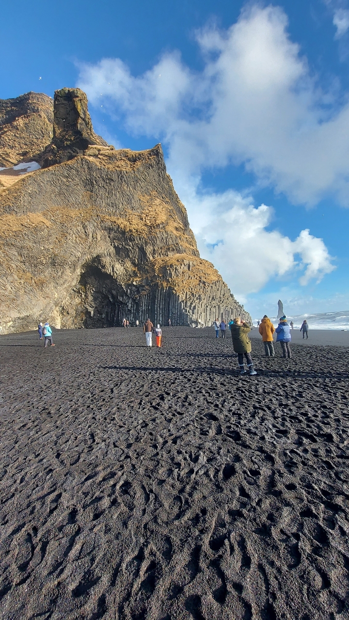 Touristes sur une plage de sable noir avec des formations de basalte colonnaire.