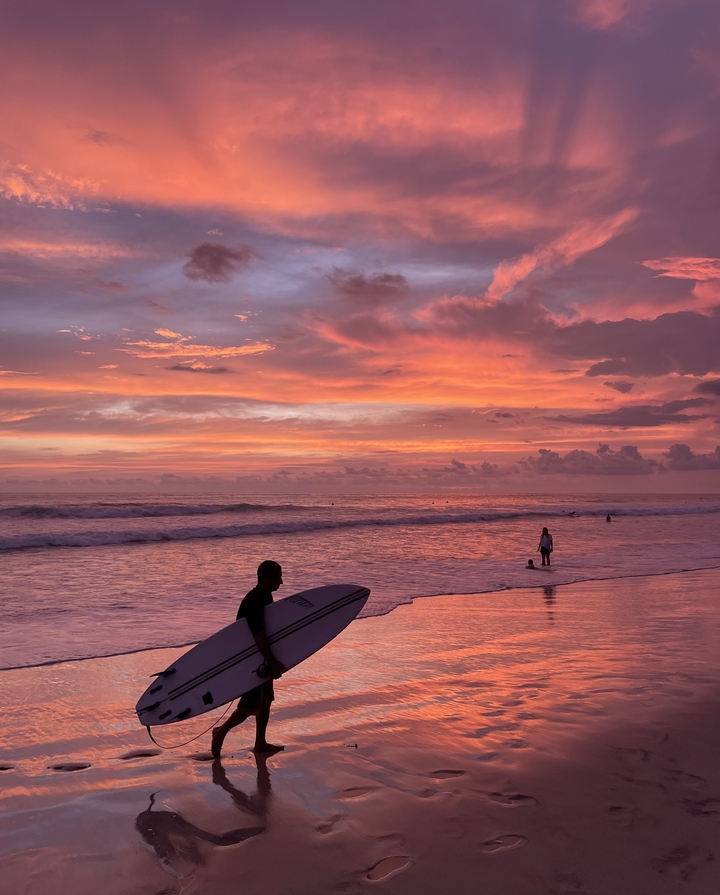 A surfer walking along the beach at a vibrant sunset.
