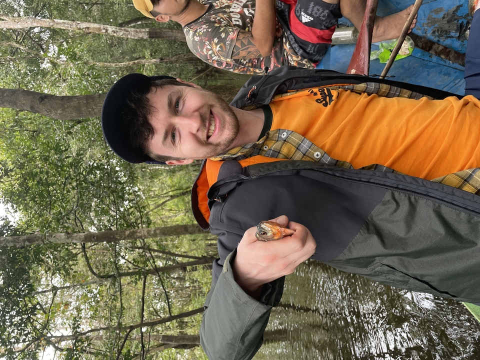 Homme en veste orange tenant un petit poisson sur un bateau.