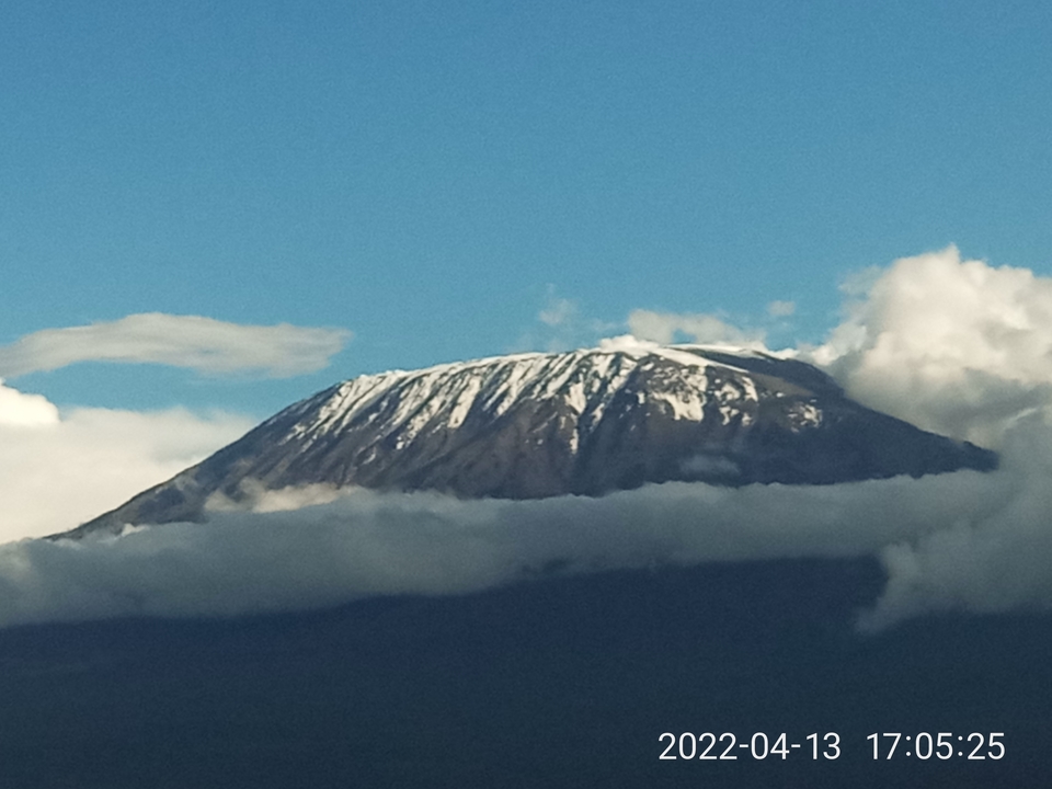 Snow-streaked Mount Kilimanjaro rises above a blanket of clouds against a blue sky.