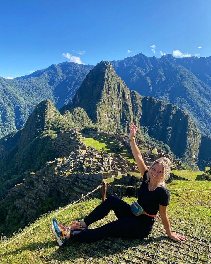 Une personne qui sourit et fait signe de la main avec le Machu Picchu en arrière-plan.