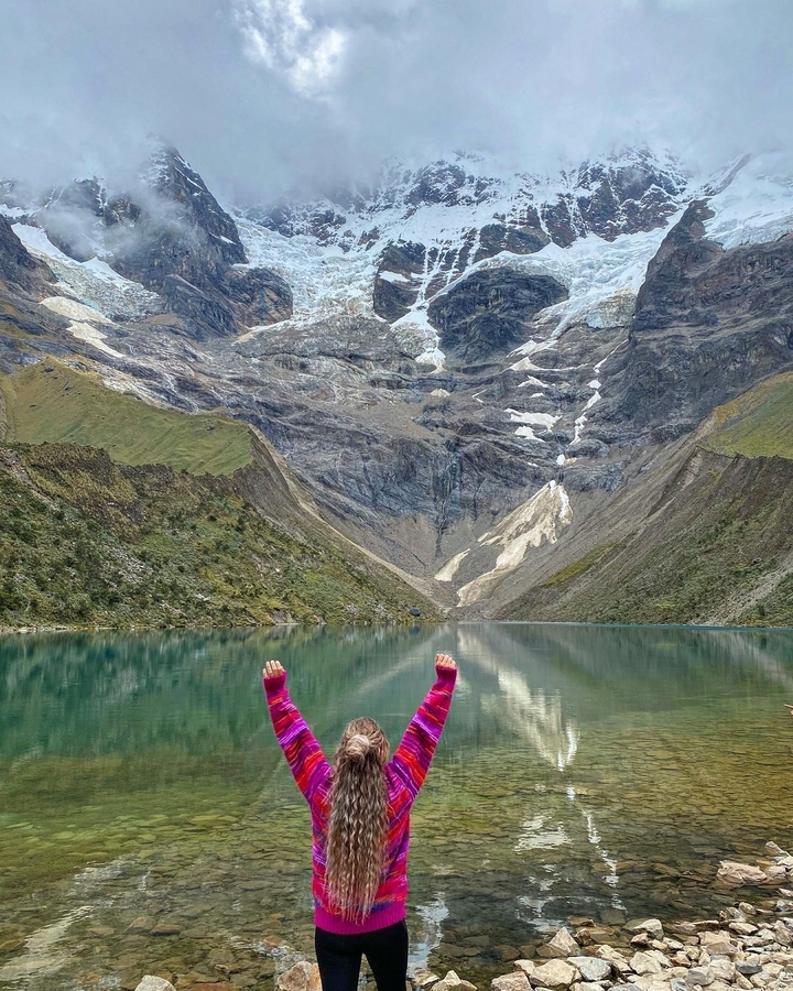 Personne avec les mains levées devant un lac aux reflets et des montagnes environnantes.