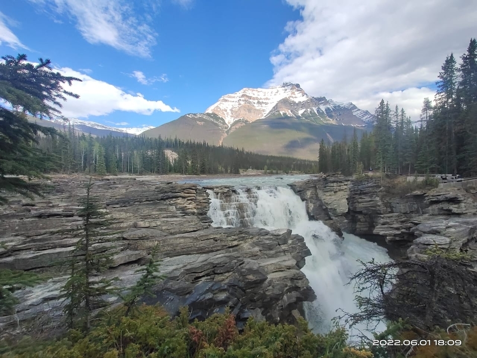 A waterfall cascades over rocks with snowy mountains in the background.