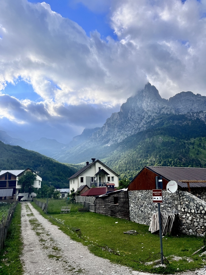 Alpine village houses beneath towering jagged peaks with dramatic sunlit clouds in the Accursed Mountains.