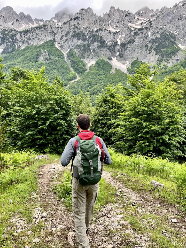 A hiker with backpack looks out over a lush green forest trail in the Albanian Alps.