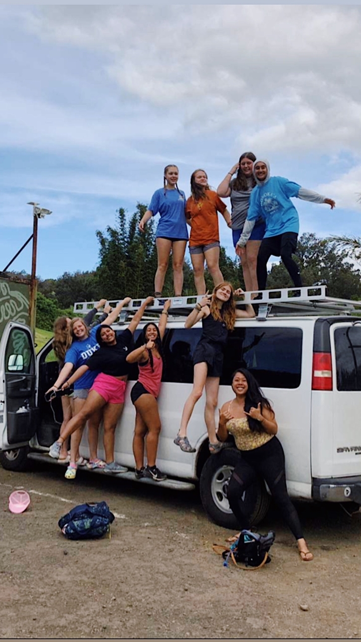 Groupe de personnes sur le toit d'un van blanc posant joyeusement à l'extérieur avec des arbres en arrière-plan.