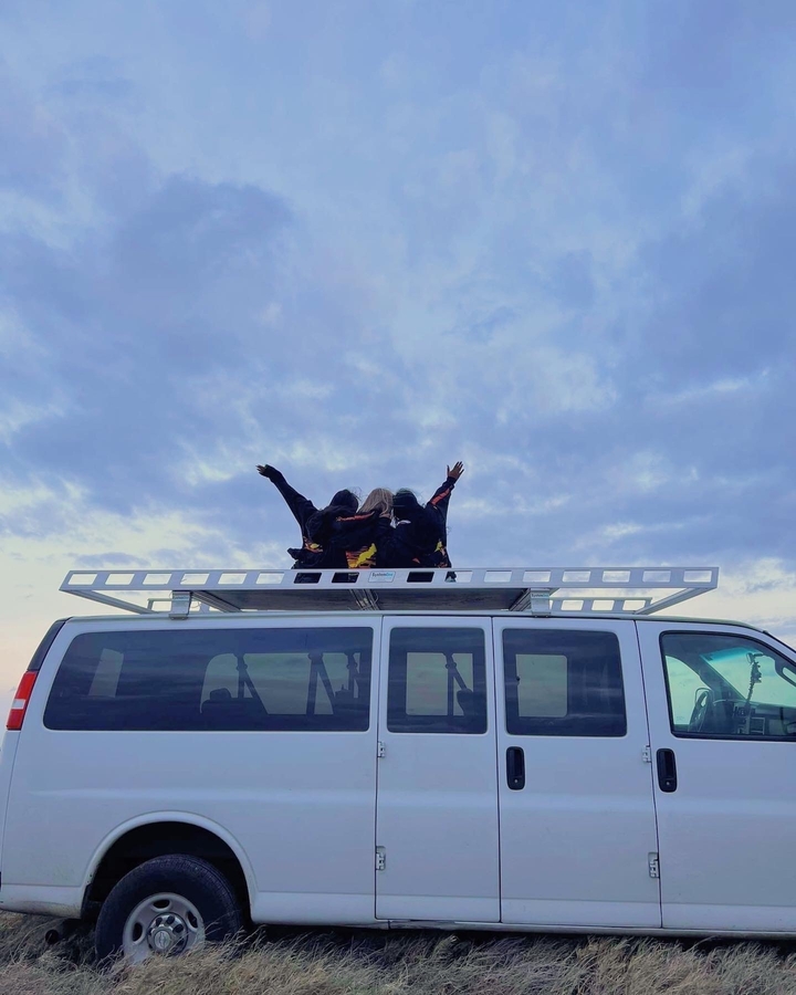Des gens assis sur le toit d'un van levant les mains vers un ciel nuageux.