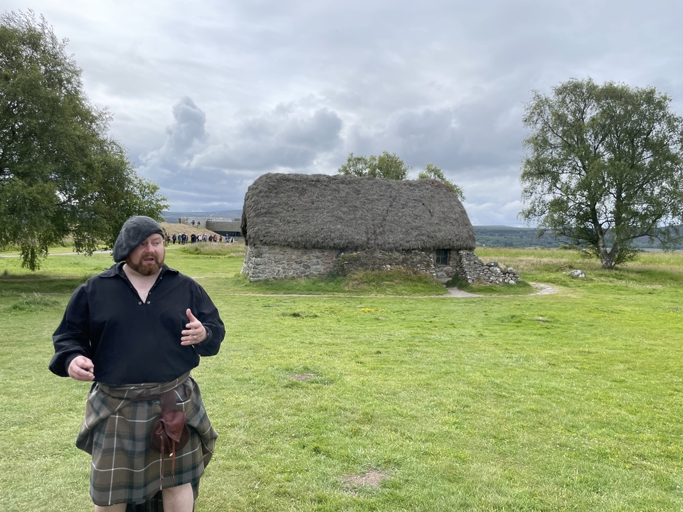 Homme en vêtements écossais parlant avec une chaumière en arrière-plan.