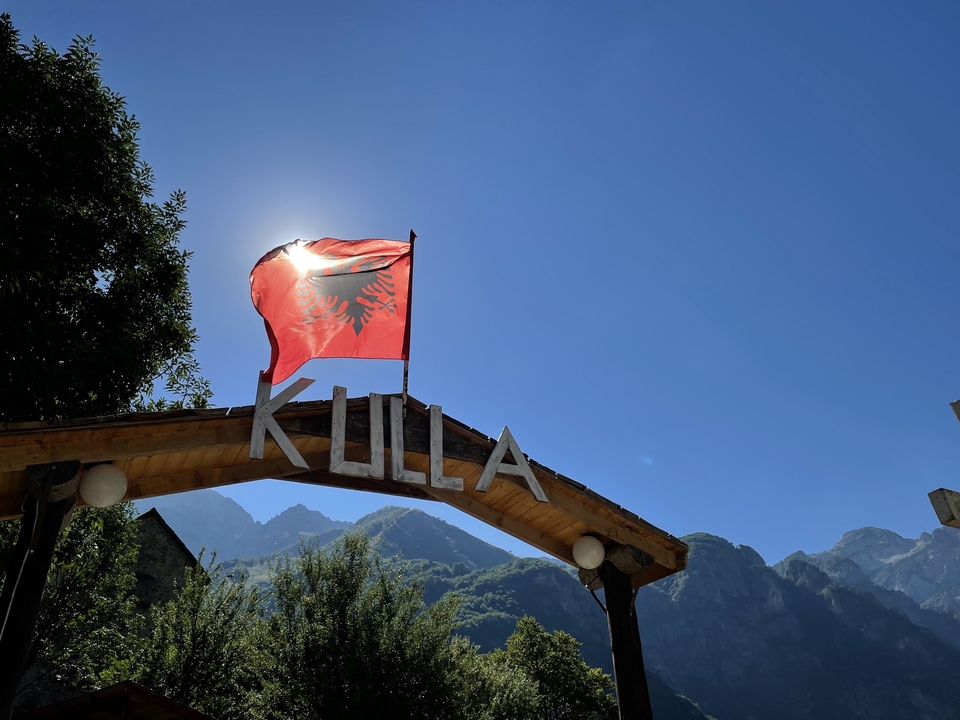 Albanian flag back-lit by the sun atop a wooden archway reading 'Kulla' against mountain backdrop