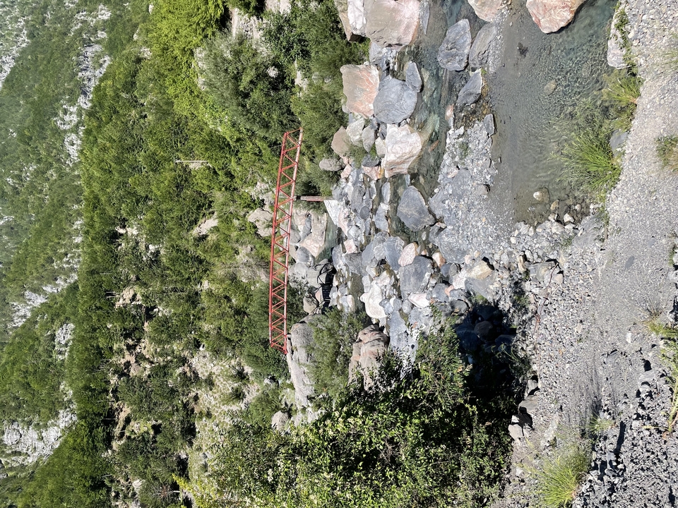Narrow red footbridge spanning a rocky mountain stream surrounded by green slopes
