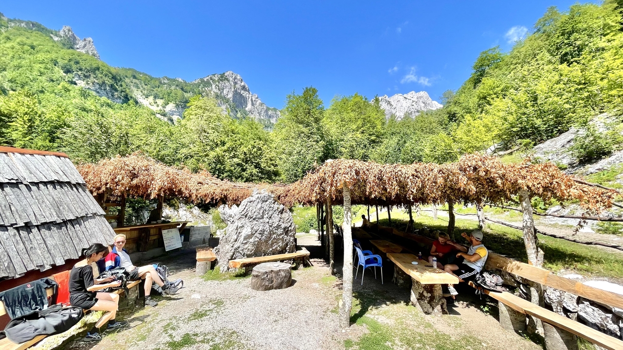 Outdoor rustic rest stop with thatched canopy, benches and hikers resting beneath green mountains