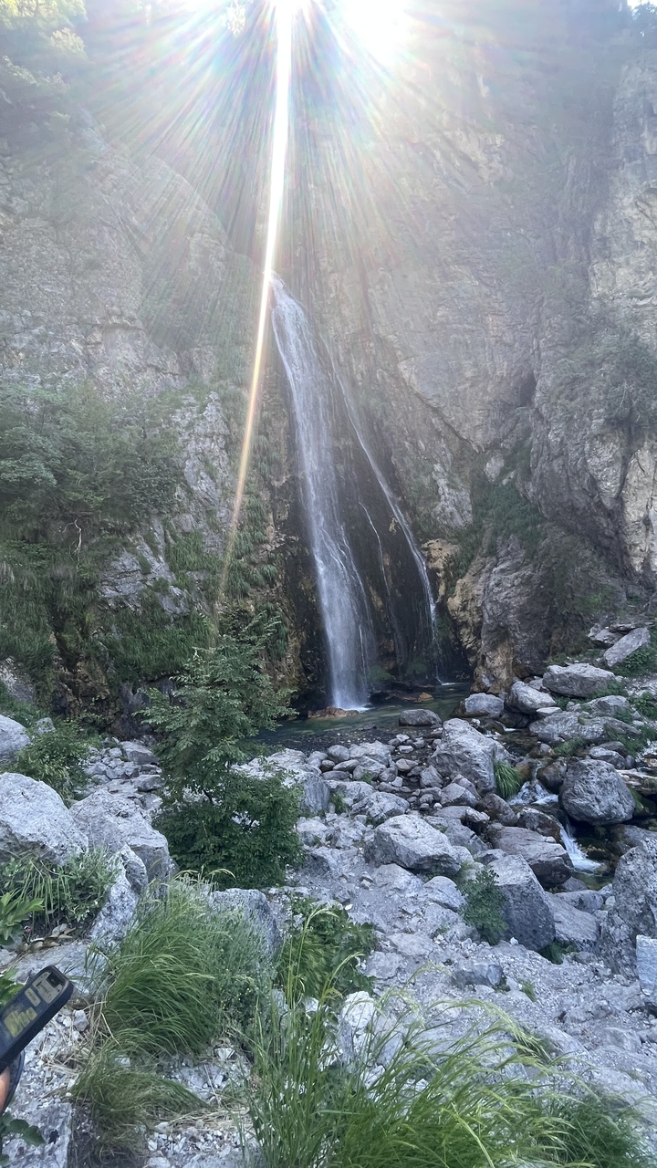 Tall narrow waterfall plunging down a rock face surrounded by green foliage with a sunbeam