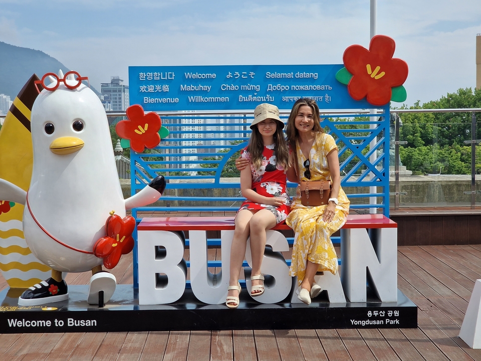 Deux personnes assises sur un panneau 'BUSAN' avec un arrière-plan urbain.