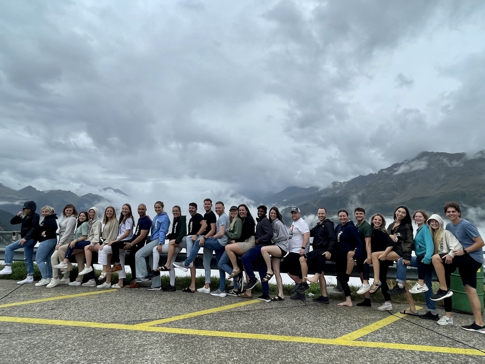 Large tour group sitting on a low wall with dramatic mountain scenery and cloudy sky