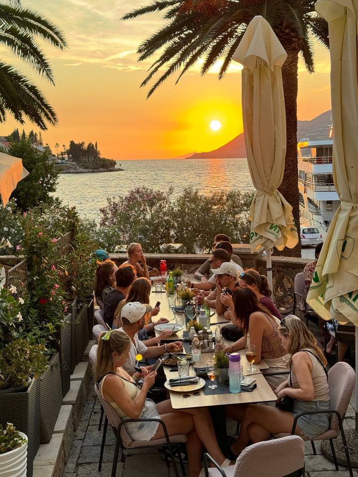Group dining outdoors with a view of the sea at sunset.