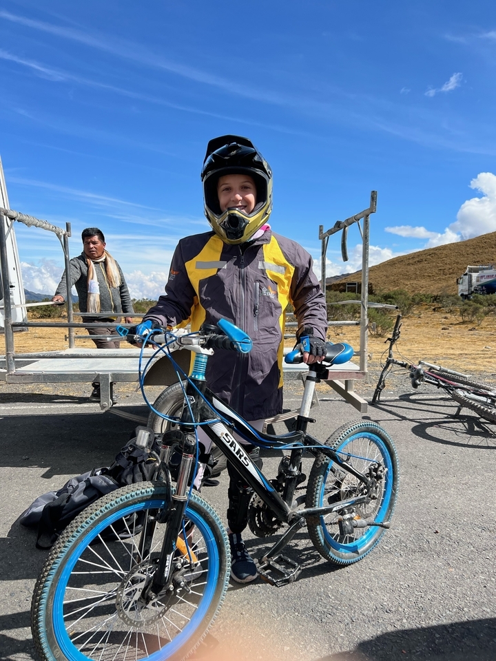 Personne portant un casque et une veste, tenant un vélo avec un paysage montagneux en arrière-plan.