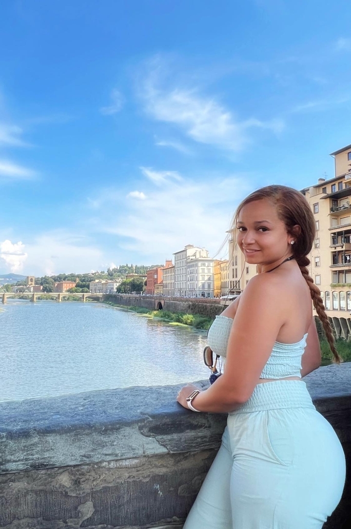 Femme souriante au bord d'une rivière avec des bâtiments et un pont en arrière-plan.