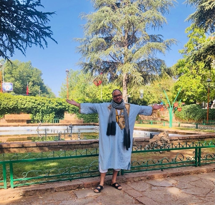 Personne en tenue traditionnelle posant dans un parc avec des arbres et une fontaine.