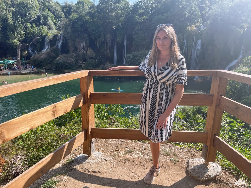 Femme près d'une rambarde en bois ; cascades et zone de baignade à l'arrière-plan.