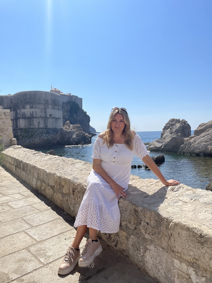 Femme en robe blanche assise au bord de la mer avec des fortifications historiques.