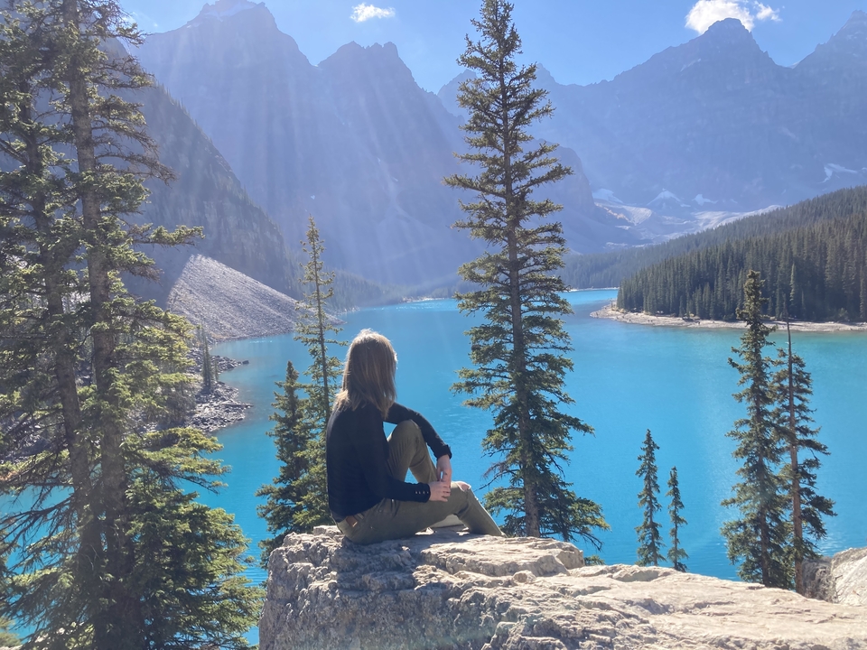 Personne assise sur un rocher surplombant un lac turquoise avec des arbres et des montagnes en arrière-plan.