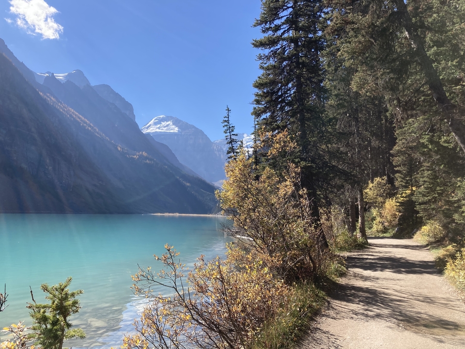 Sentier le long d'un lac turquoise avec une montagne enneigée au loin.