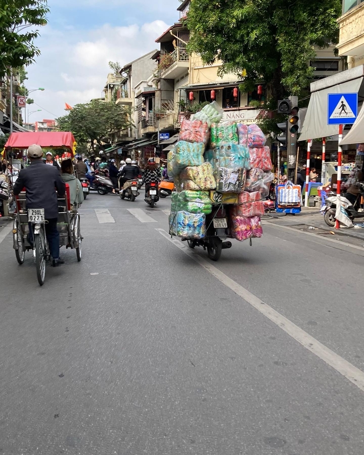 Scène de rue animée avec des gens et des véhicules, y compris une moto chargée de marchandises.