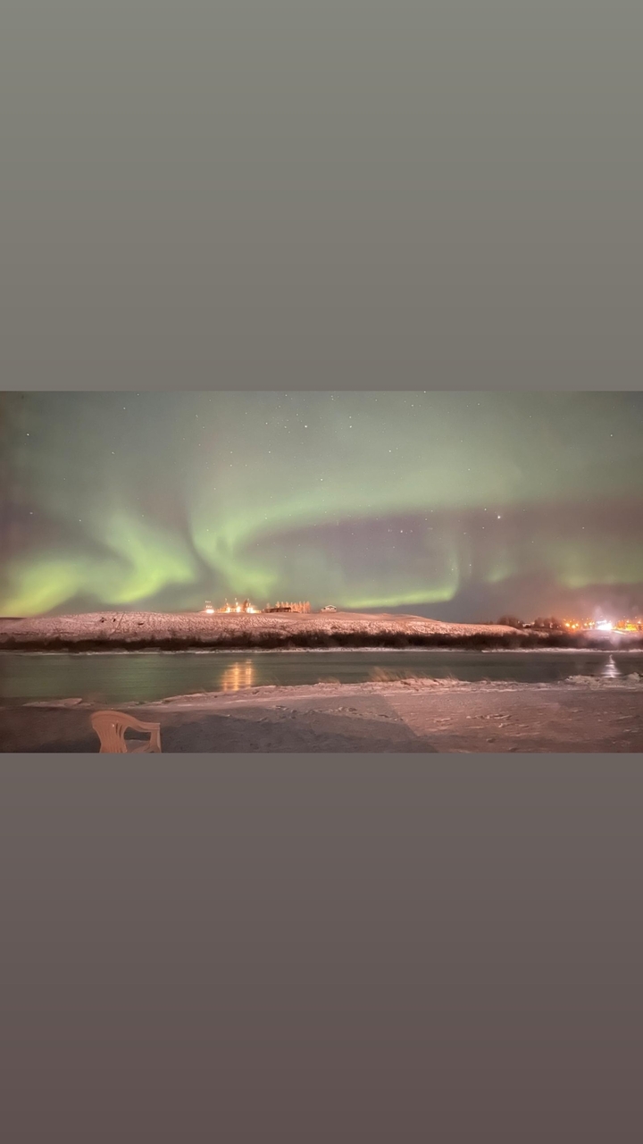 Aurores boréales au-dessus d'un paysage enneigé avec des lumières au loin.