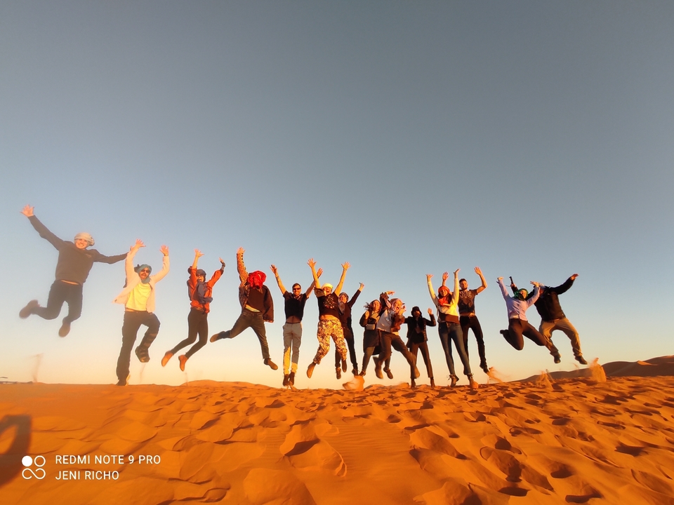 A group of people jumping off a sand dune with the sun setting in the background.