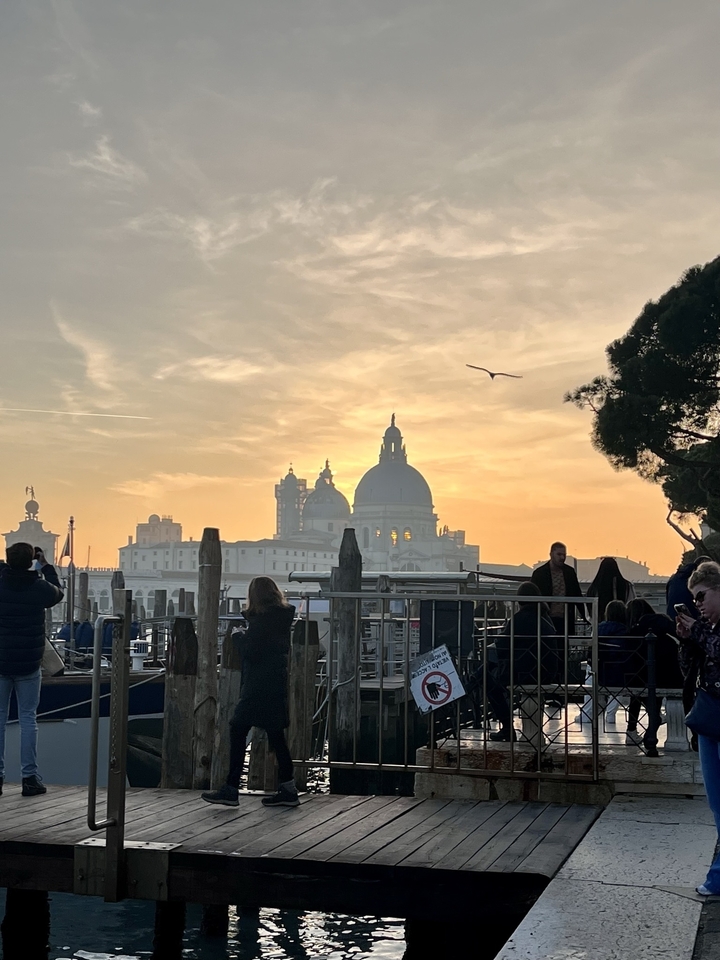 Silhouette d'un bâtiment avec des dômes contre un ciel de coucher de soleil, avec des personnes et des arbres au premier plan.