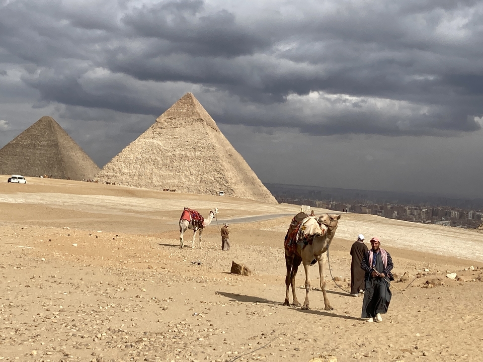 People and camels in front of the pyramids under a cloudy sky.
