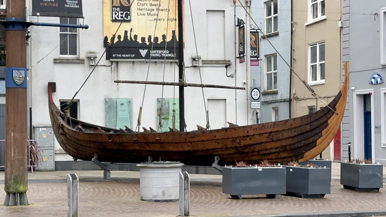 Un bateau en bois de style viking exposé sur une place de ville avec des bâtiments en arrière-plan.