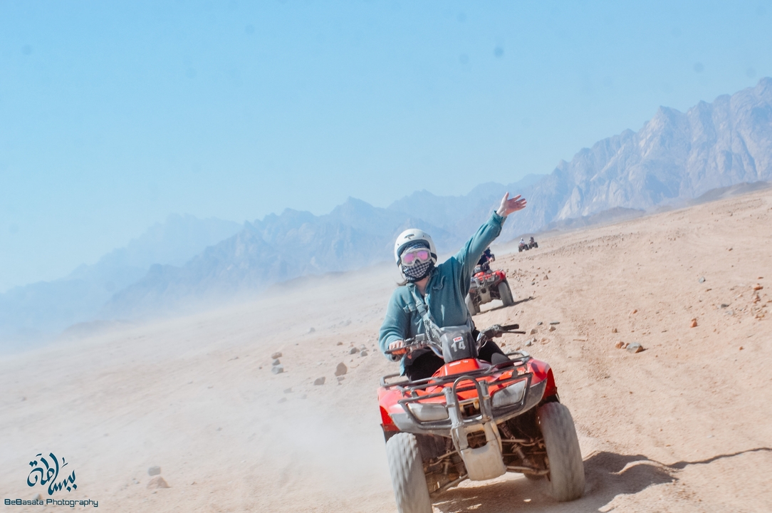 Des gens qui font du quad sur un désert sablonneux avec des montagnes en arrière-plan.