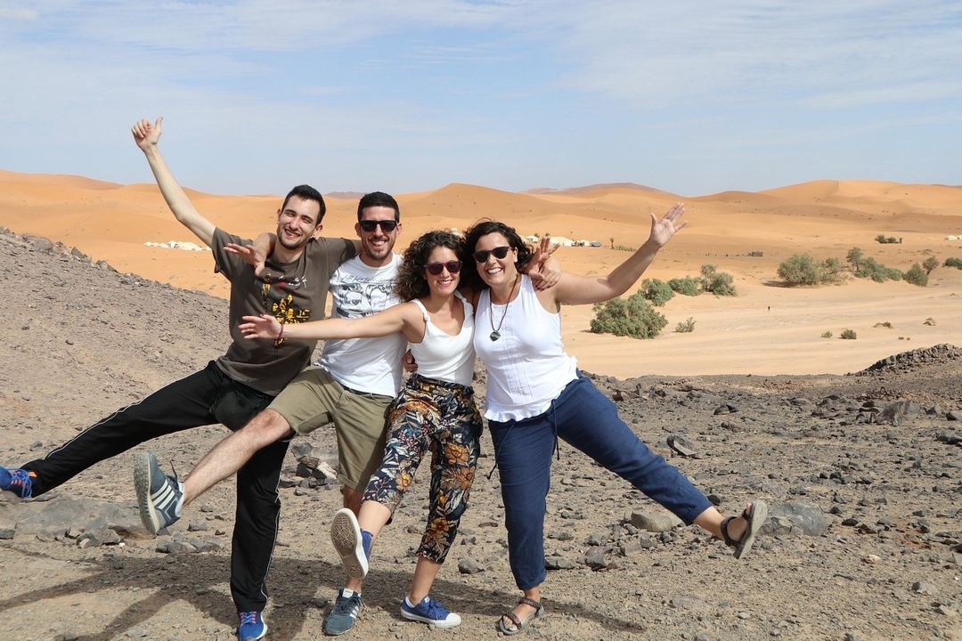 A group of four people posing energetically in a desert with sand dunes in the background.