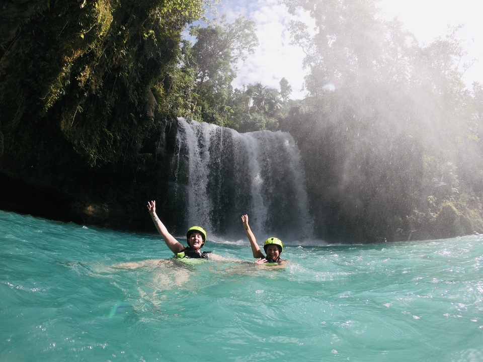 Two people swimming in a turquoise water in front of a waterfall.